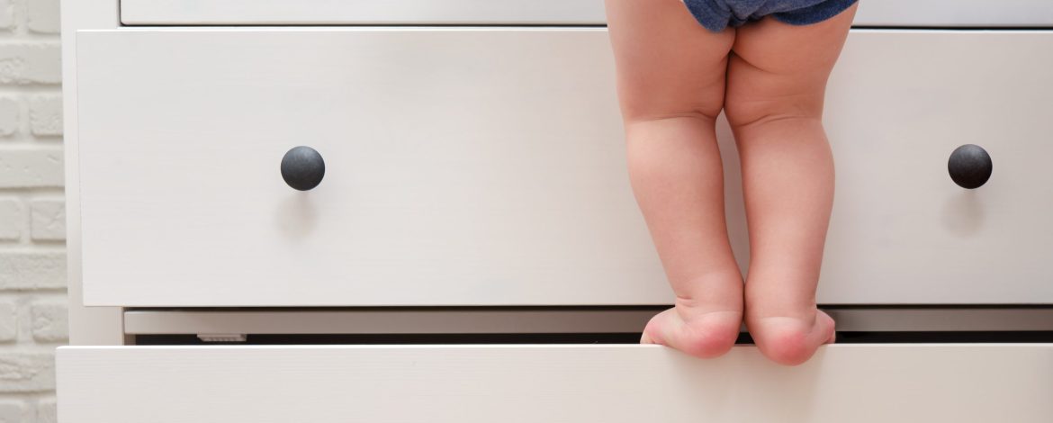 Toddler baby climbed up on the open chest of drawers. Child boy stood on a tall drawer of a white cabinet. Kid age one year